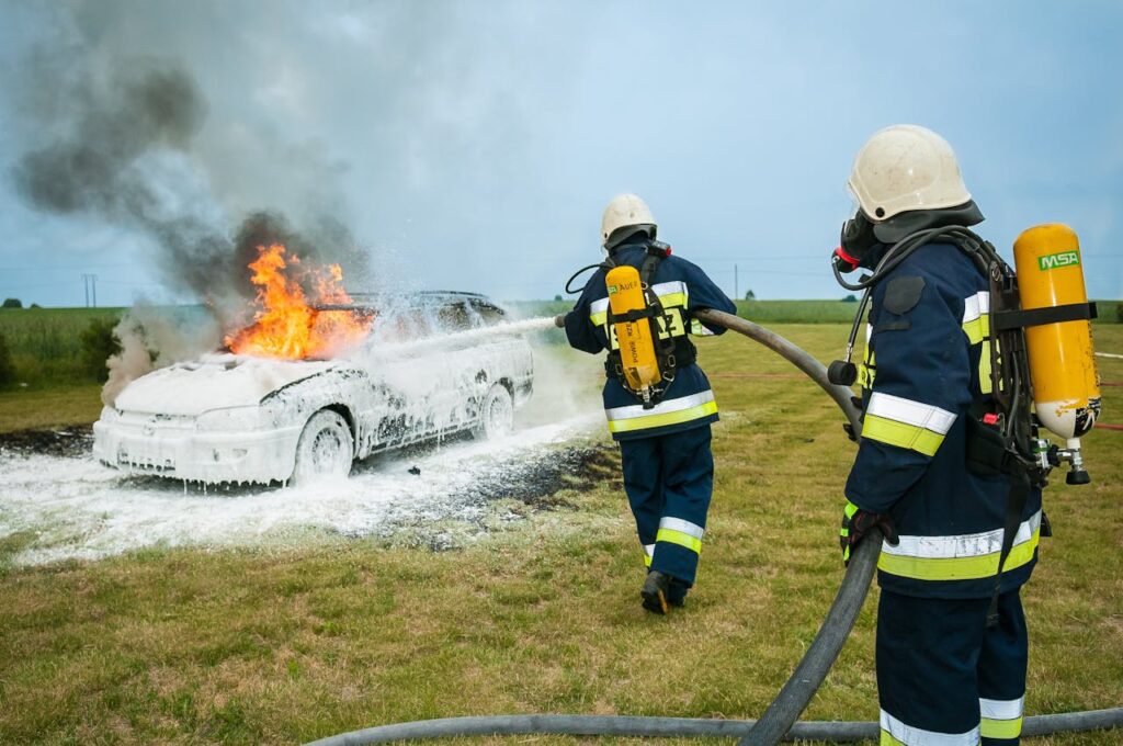 Firefighters tackle a blazing car using foam to extinguish the flames in an outdoor setting.