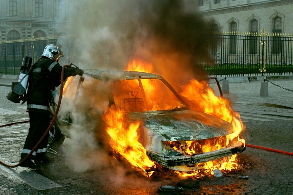 Firefighter in protective gear extinguishes a burning car in a city street.
